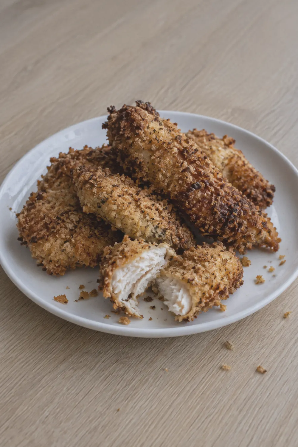 Air fryer chicken tenders piled on a white plate with uneven golden-brown breading and a few breadcrumb crumbs scattered around
