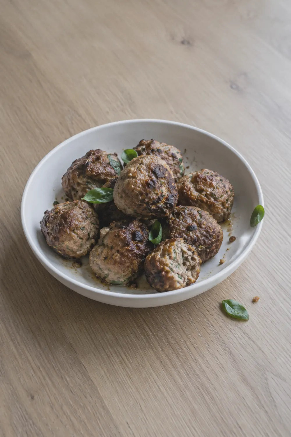 Browned air fryer meatballs in a shallow white bowl on a pale oak surface with a few basil leaves scattered nearby