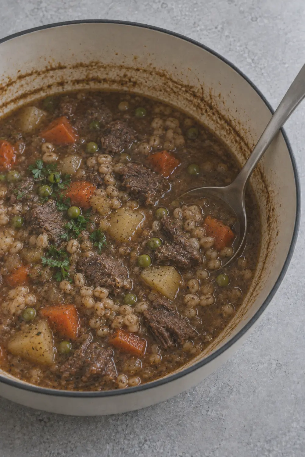Finished beef barley soup in a Dutch oven with bright green peas scattered throughout the thick brown broth alongside tender beef chunks, barley, and vegetables with fresh parsley on top