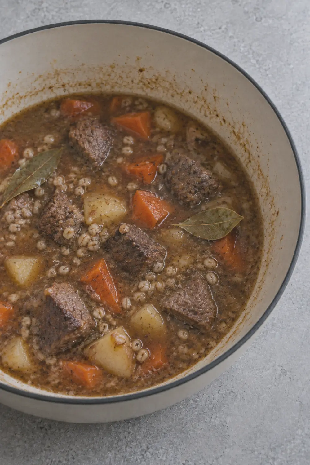 A Dutch oven filled with simmering beef barley soup showing chunks of beef, plump barley, diced carrots, and potatoes in a rich dark brown broth with thyme sprigs visible