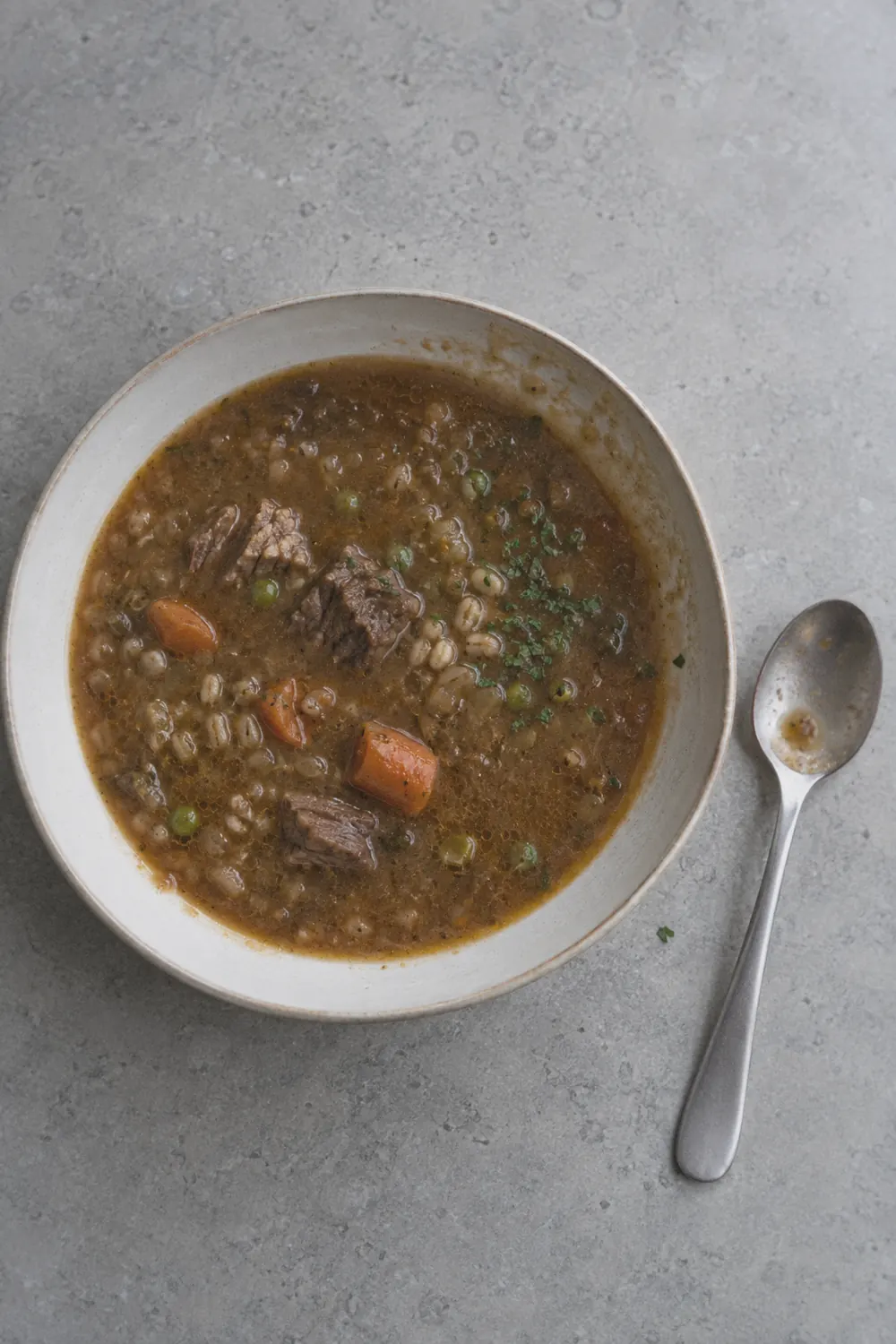 A large bowl of beef barley soup with tender chunks of beef, pearl barley, carrots, celery, and potatoes in a rich brown broth on a wooden table with crusty bread and a linen napkin alongside.