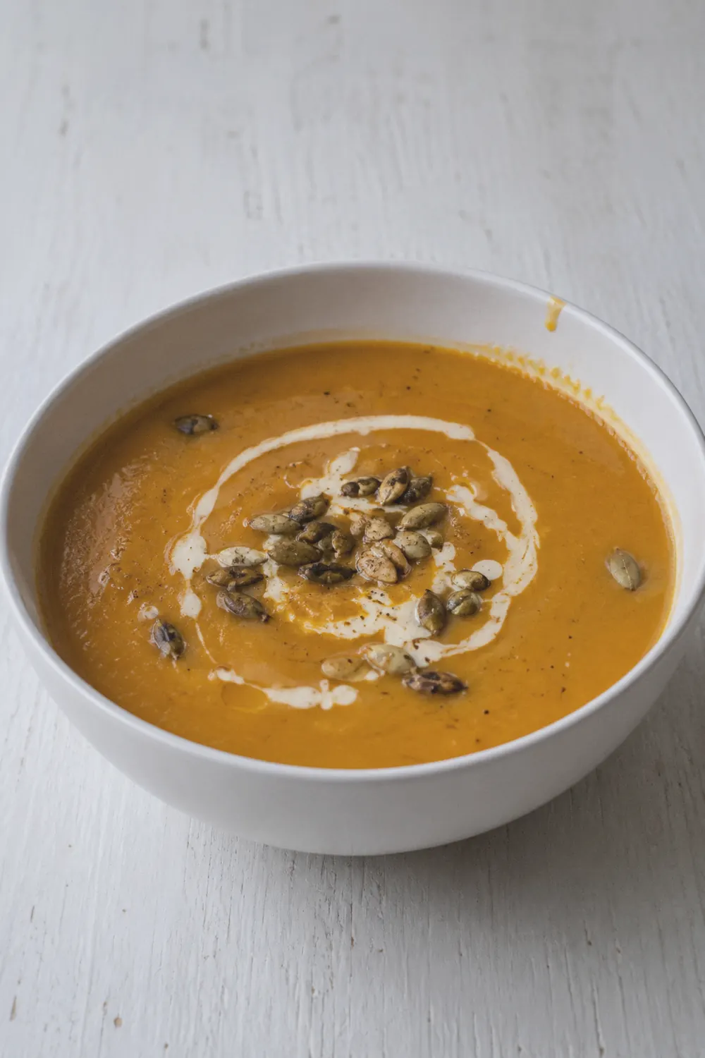 A bowl of smooth butternut squash soup with a swirl of cream and a few toasted pumpkin seeds on top, set on a white painted wood surface