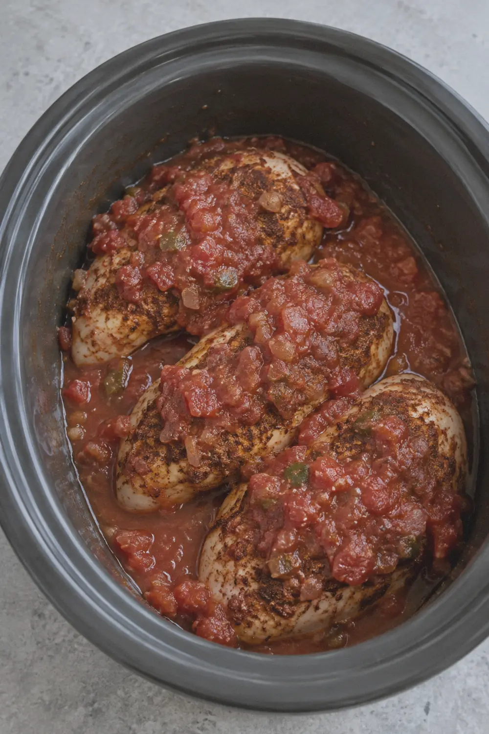 Boneless skinless chicken breasts seasoned with a dark spice rub sitting in a single layer inside a slow cooker insert with chunky salsa spooned over the top