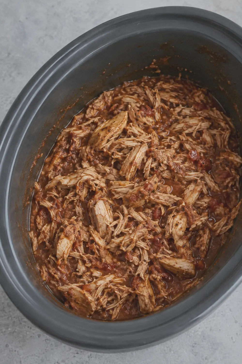Shredded chicken tossed back into the slow cooker insert with the reduced salsa cooking liquid, showing irregular strands and chunks of meat coated in sauce with juices smeared on the inner rim