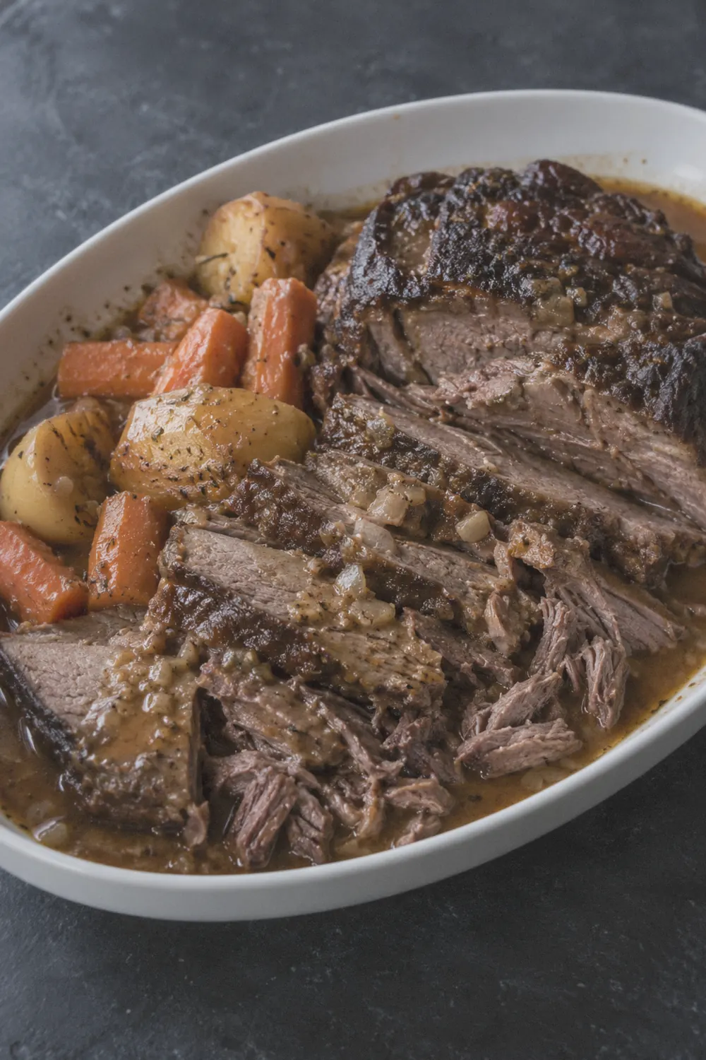 A sliced crockpot pot roast on a dark slate surface with potatoes and carrots, rich brown gravy pooled around the meat