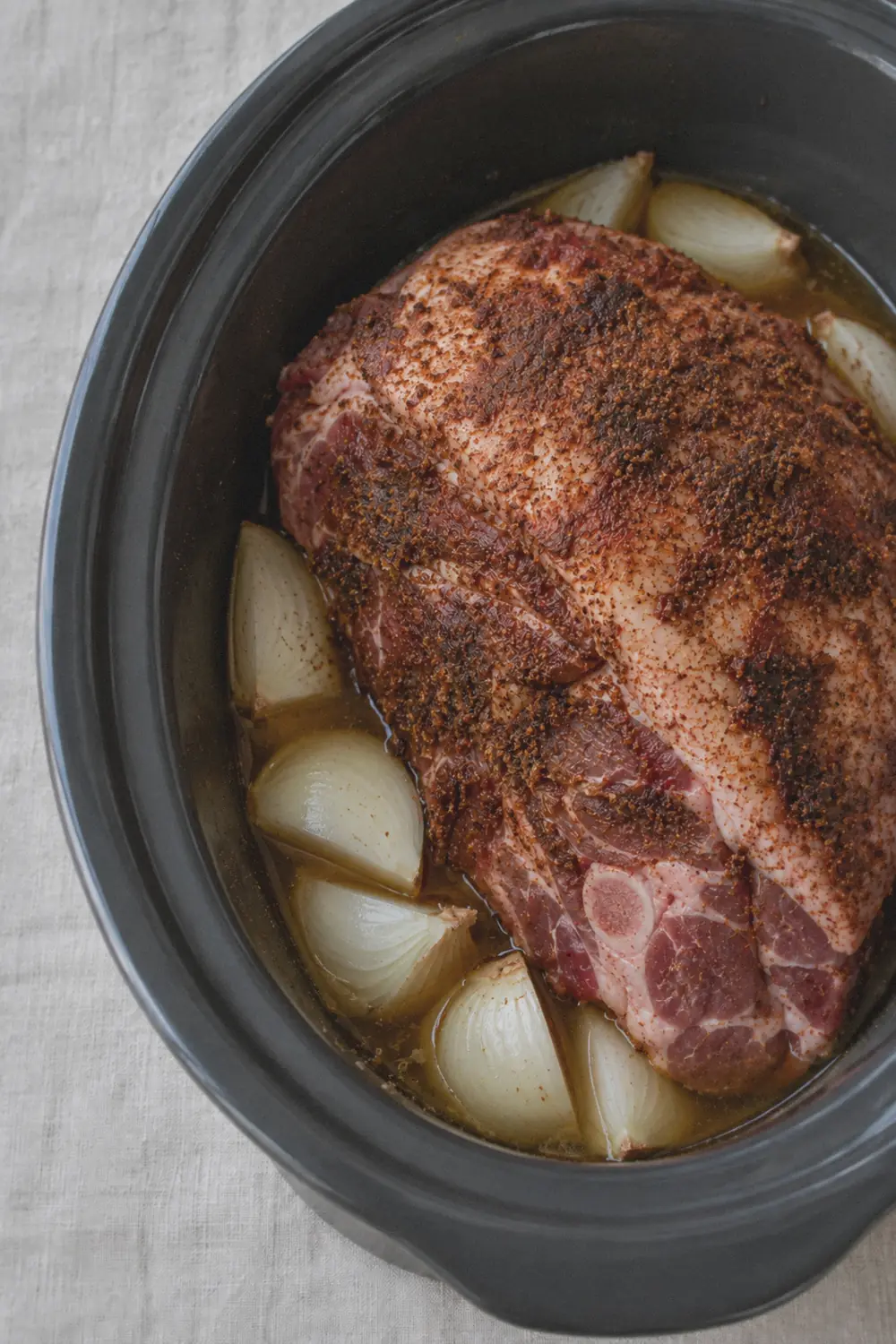 Pork shoulder inside a slow cooker insert with visible dry rub on the surface and onion quarters underneath, raw chicken broth visible around the sides