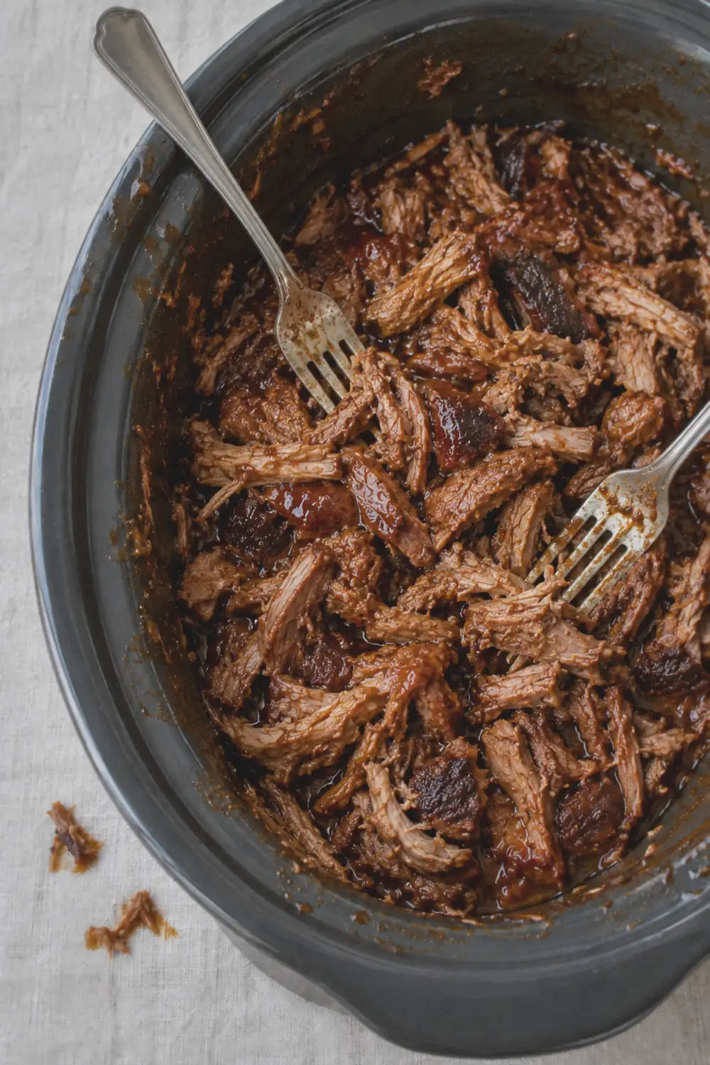 Shredded pulled pork tossed with barbecue sauce in a slow cooker insert, showing a mix of strands, flakes and larger chunks with sauce smeared on the inner rim