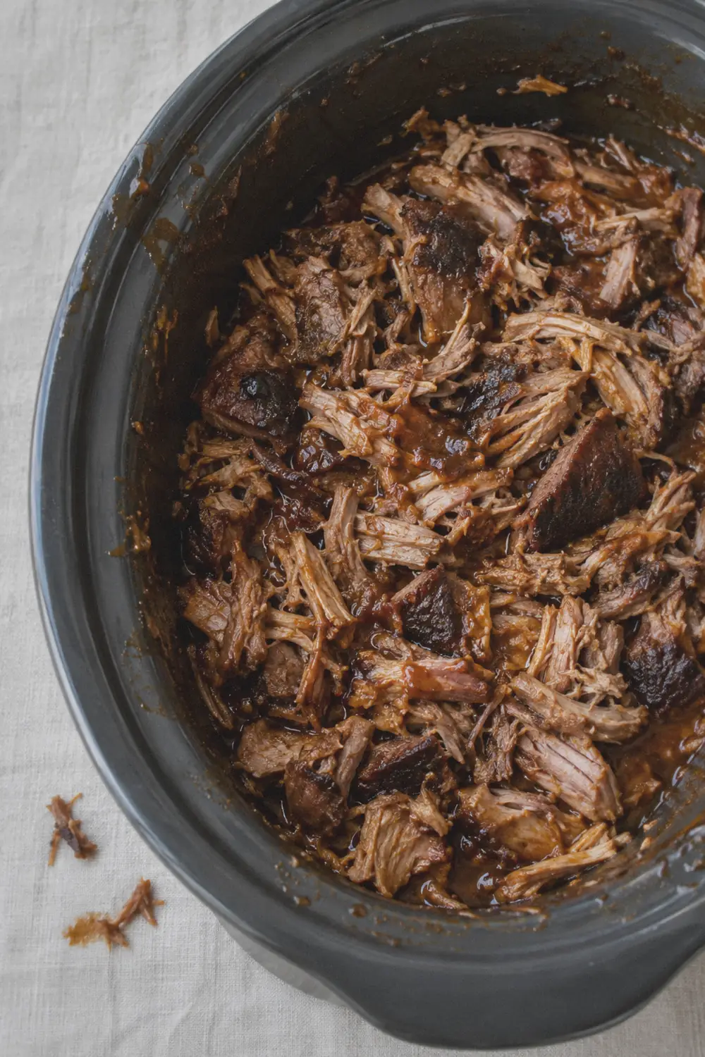 Tender crockpot pulled pork shredded with two forks in a slow cooker insert, showing irregular strands and chunks of juicy meat on a light beige linen surface.