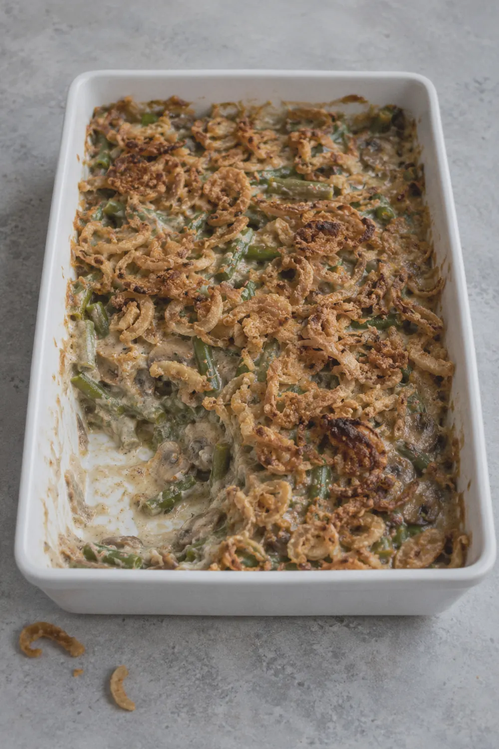 Green bean casserole in a white ceramic baking dish topped with crispy fried onions on a light grey stone surface
