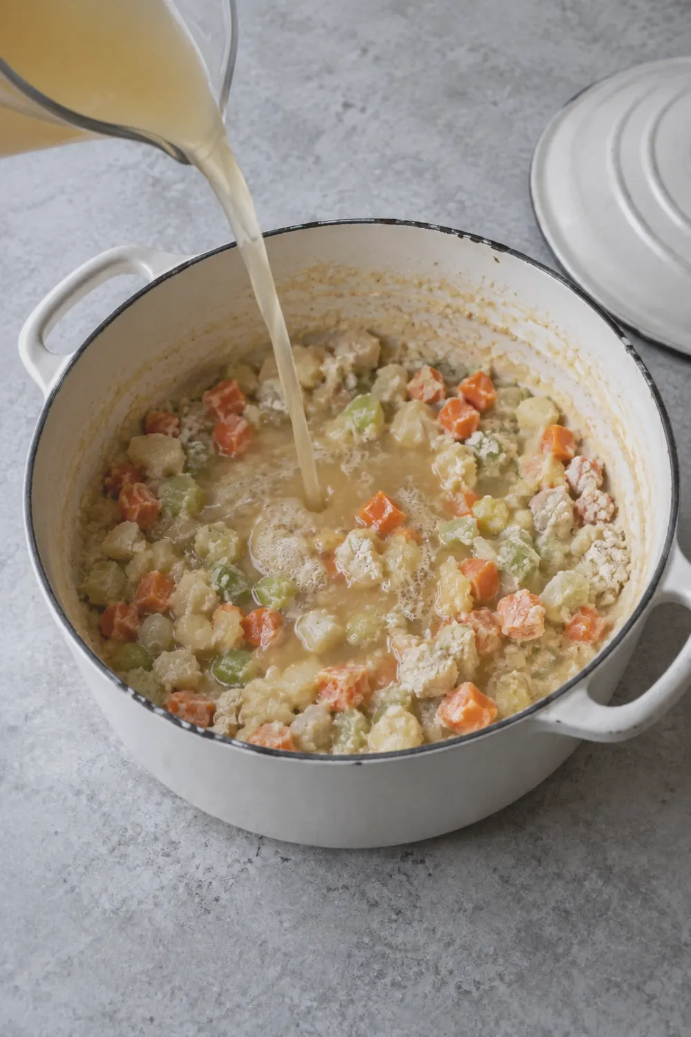 A Dutch oven with diced onion, carrots, and celery coated in a flour-butter roux, with chicken broth being poured in and the mixture starting to thicken