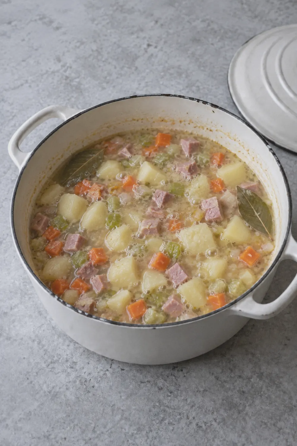 Ham and potato soup simmering in a Dutch oven with chunks of pink ham and potato cubes visible in a pale broth with carrots and celery pieces throughout