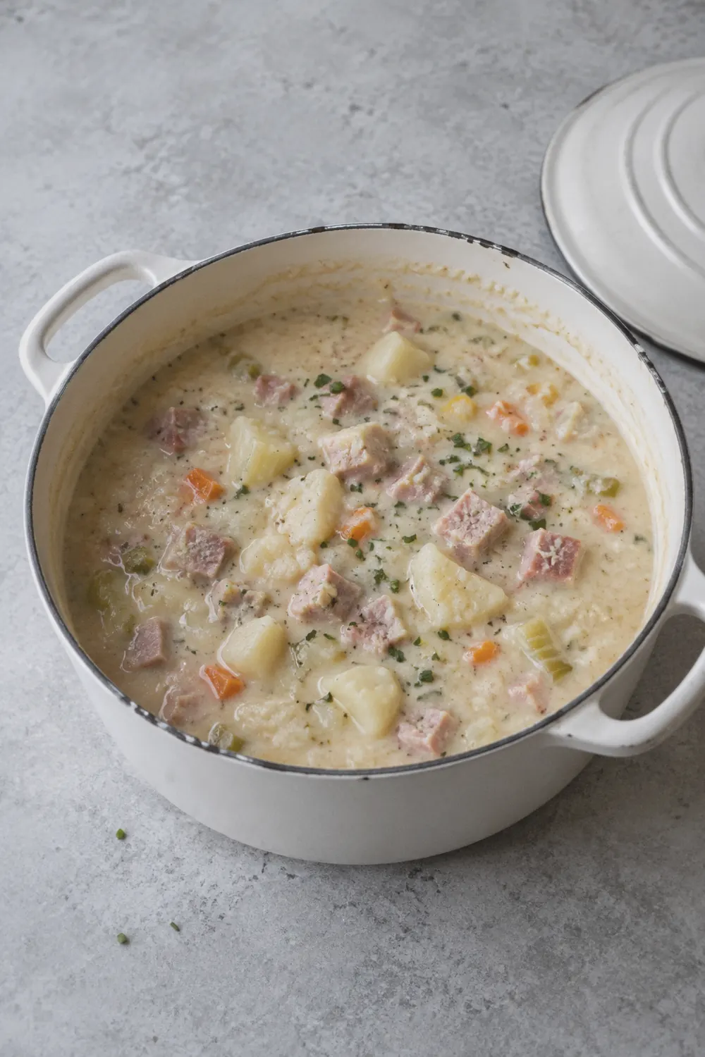 A bowl of creamy ham and potato soup with chunks of pink ham and soft potato pieces in a thick pale broth topped with chopped chives on a light concrete surface