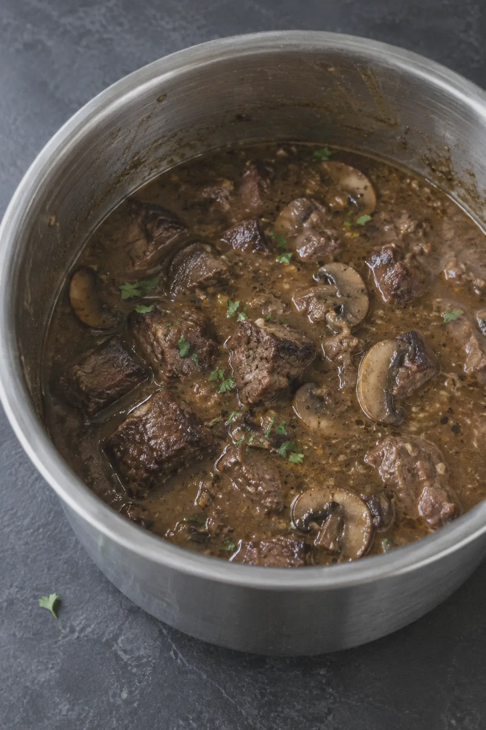 A stainless steel Instant Pot insert filled with tender beef tips in thick brown mushroom gravy, on a dark slate surface