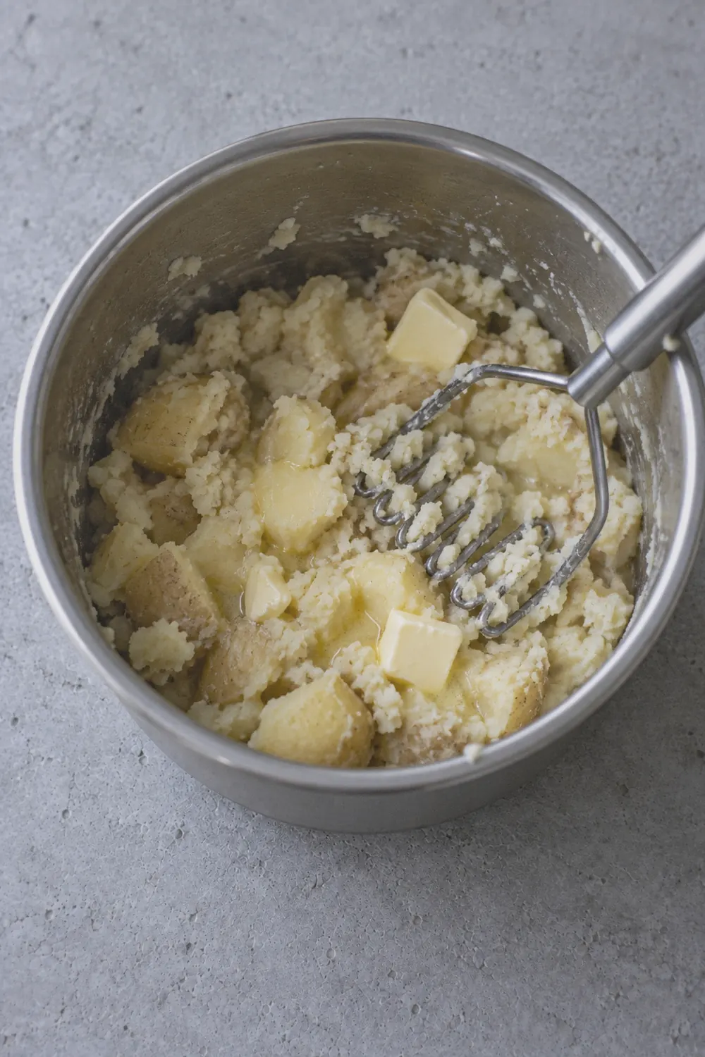 Mashed potatoes being worked with a potato masher in the Instant Pot inner pot, butter partially melted and streaked through the potatoes, the texture chunky and uneven mid-mash