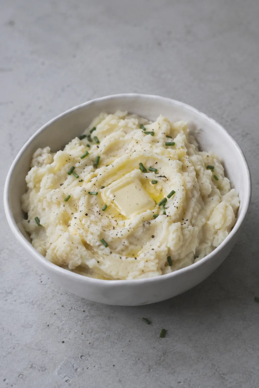 A generous serving of creamy mashed potatoes in a round white bowl on a light concrete surface, with a pat of butter melting into the center and a few flecks of black pepper and chopped chives scattered unevenly on top