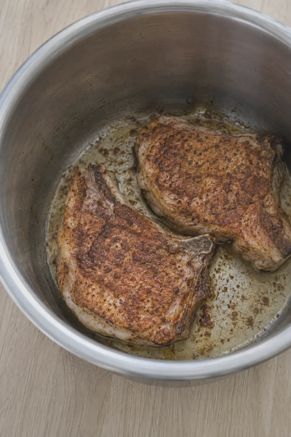 Two pork chops searing in an Instant Pot inner pot with oil, showing a golden-brown crust forming on the top side while the bottom side presses against the hot surface, sliced onions waiting beside the pot