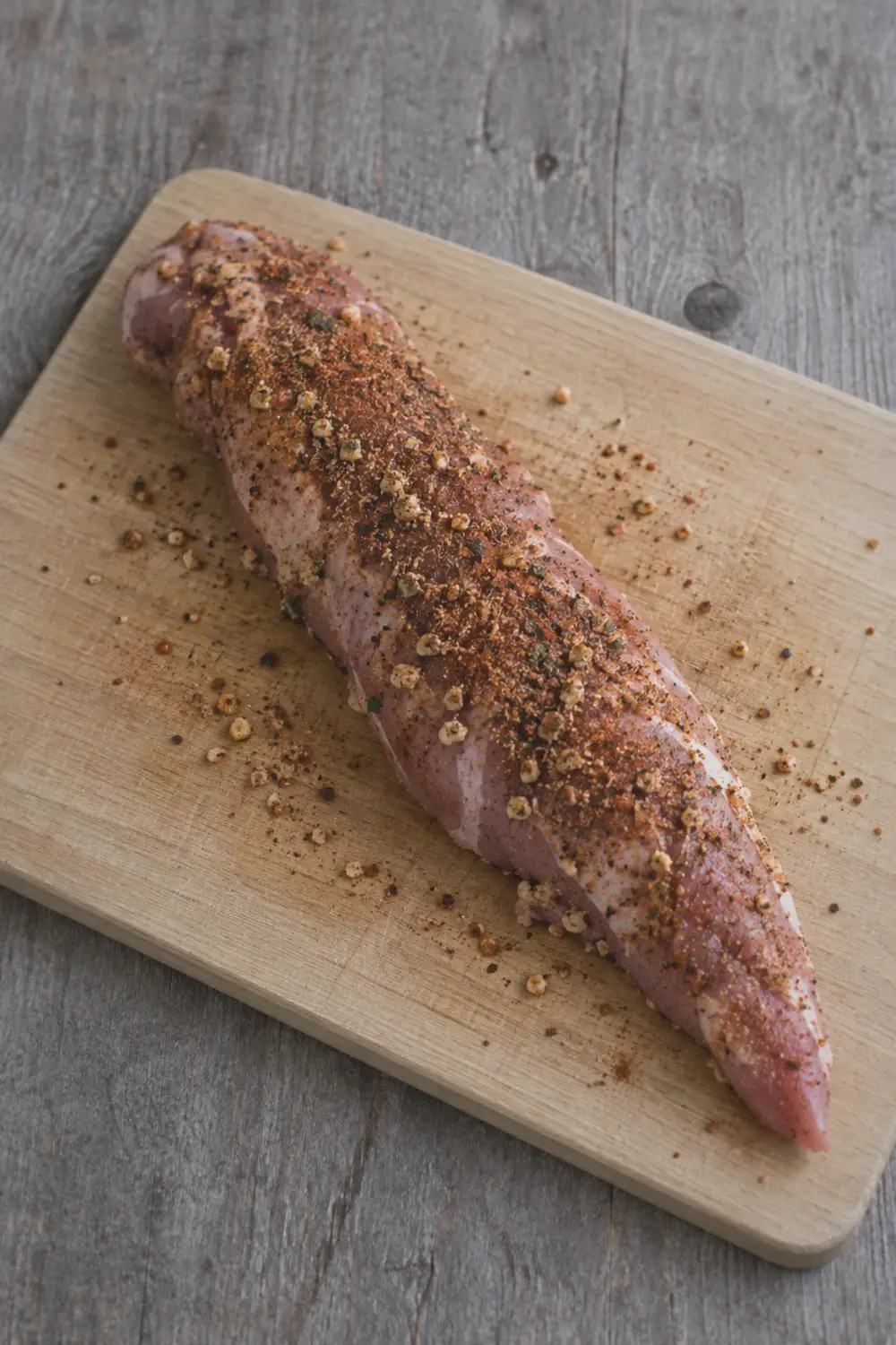 Pork tenderloin rubbed with spices resting on a cutting board before searing, showing the even layer of garlic-paprika seasoning pressed into the surface of the raw meat