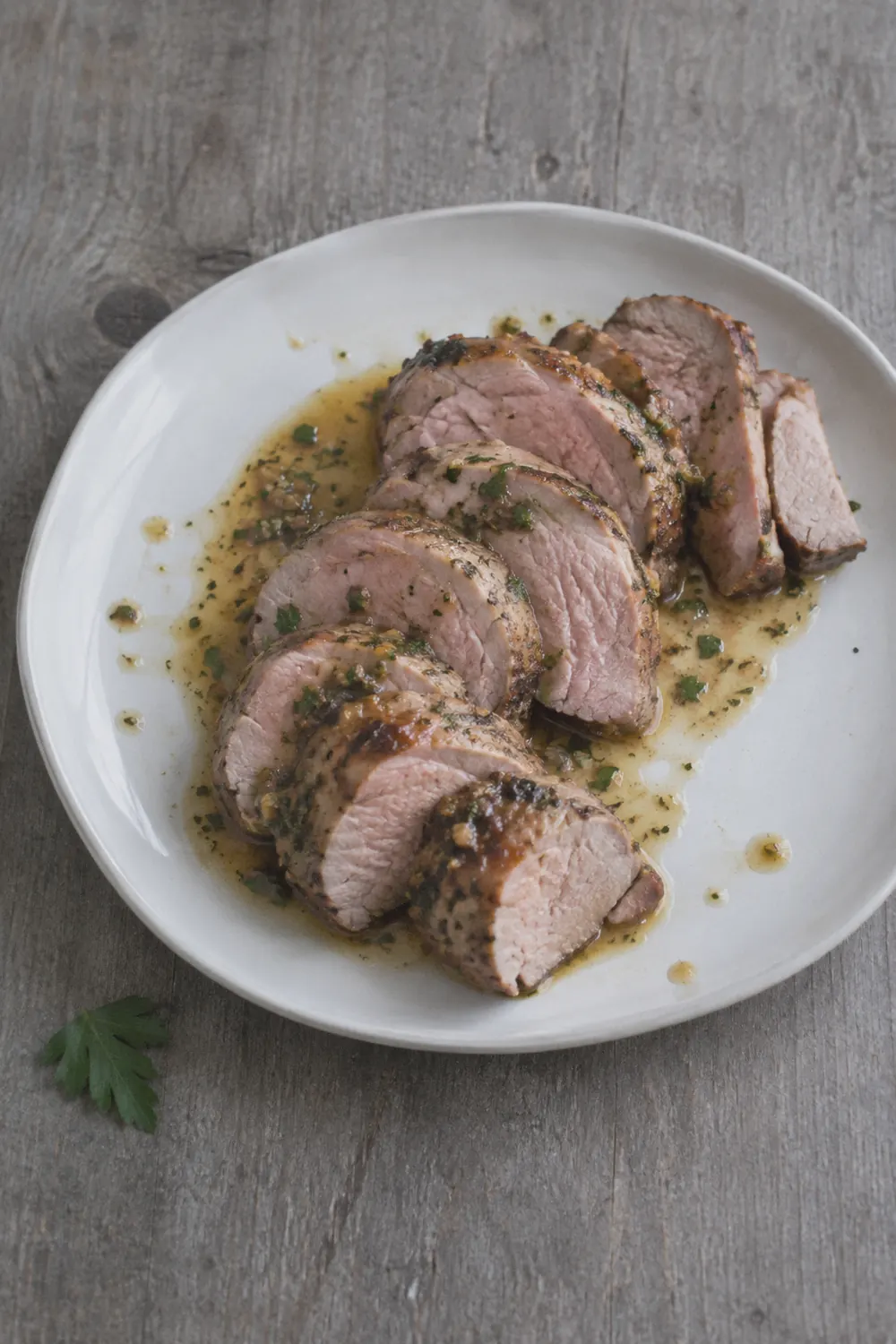 Sliced pork tenderloin fanned on a white plate with a thin herb pan sauce pooled around the slices, a few drops of sauce on the plate rim, on a weathered grey wood surface