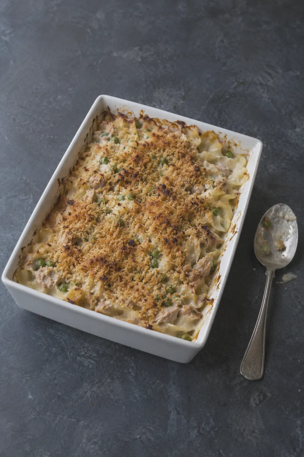 Tuna noodle casserole in a 9x13 baking dish with golden breadcrumb topping and peas visible throughout the creamy noodle filling